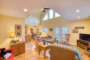 A spacious living room with a high ceiling, a fireplace, a television, and a dining area with a kitchen in the background. - Carr House Upper (Lake Junaluska)