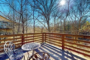 Eine Holzterrasse mit Metallstühlen und einem Tisch, umgeben von Bäumen und blauem Himmel.