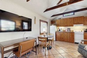 A compact kitchen with wooden cabinets, a dining table with chairs, a TV mounted on the wall, and a window with blinds. - Charming Brevard cottage perfect for a getaway with local shops & breweries! (Brevard)