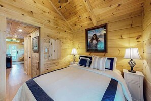 A wooden-paneled room with a bed, bedside lamps, a framed picture on the wall, and a door leading to another room. - Scenic Cabin w/ Private Hot Tub, Amazing Views, & Covered Porch. Nearby Skiing (Mars Hill)