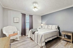 Ein Schlafzimmer mit Bett, Sessel, Nachttisch, einem Fenster mit Vorhängen und einer Deckenleuchte.