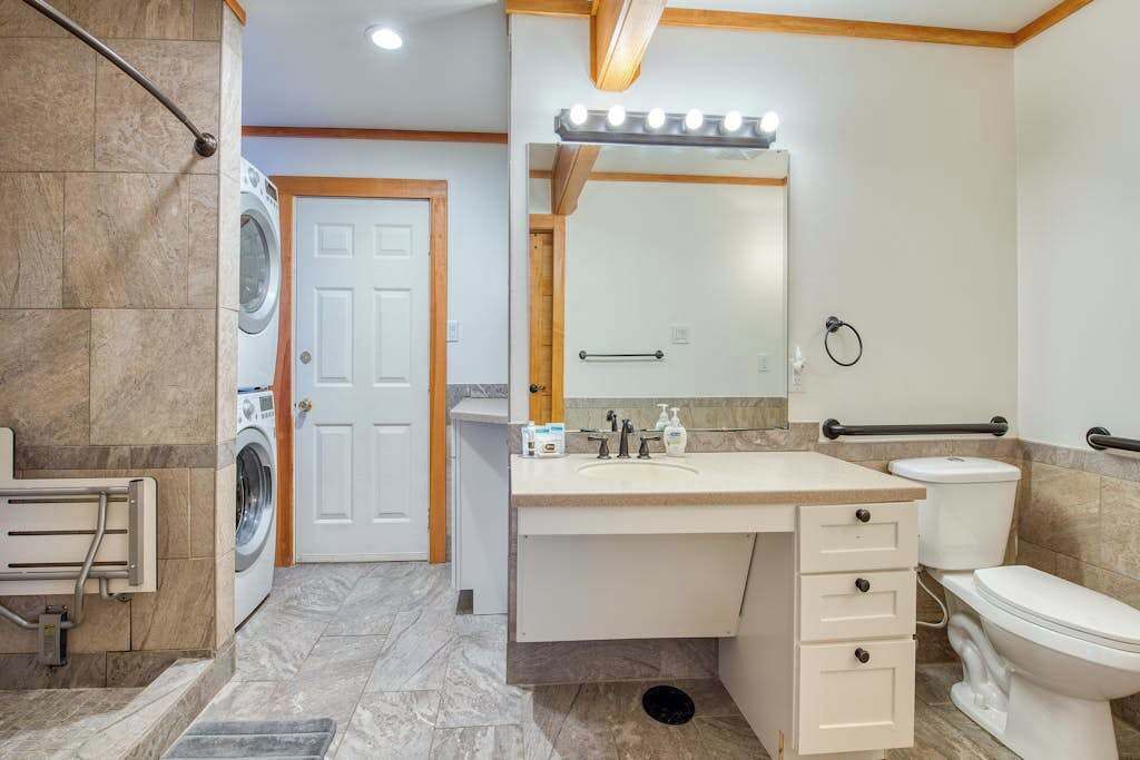 Une salle de bain équipée d’un lave-linge, d’un évier, d’un miroir avec éclairage, de toilettes et d’une barre d’appui.