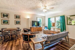 Ein Wohnzimmer mit einem Holzesstisch, Stühlen, einer Couch, einem Fernseher und gerahmten Bildern an den Wänden.