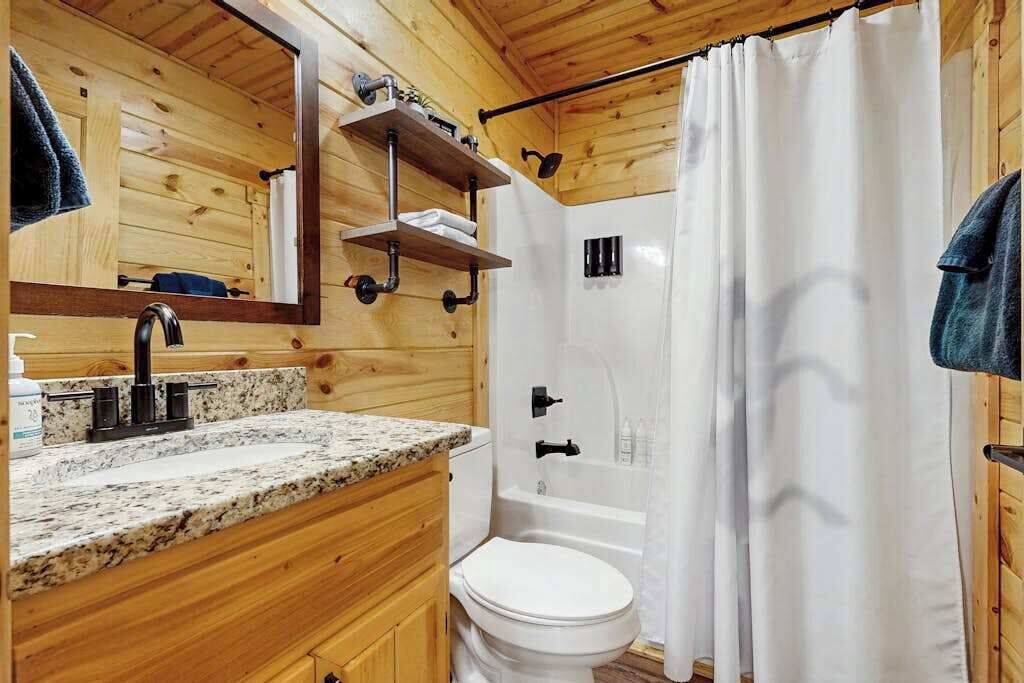 A wooden cabin bathroom with a granite countertop sink, a white toilet, a bathtub, and a shower curtain.