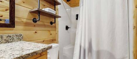 A wooden cabin bathroom with a granite countertop, a toilet, a shower with a curtain, and a shelf with a towel and a sign.