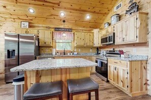 A cabin-style kitchen with wooden cabinets, granite countertops, stainless steel appliances, and a central island with bar stools. - The Shepherd’s House (Murphy)