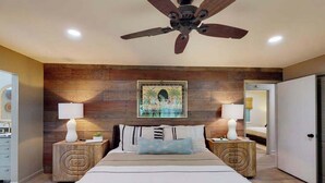 Quarto com cama grande, cabeceira de madeira, duas mesas de cabeceira com abajures, ventoinha no teto e uma moldura com imagem na parede.