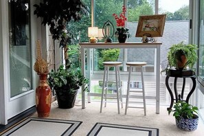 Ein Zimmer mit Blick durch große Fenster, ein kleiner Tisch mit Lampe, zwei Hocker und Topfpflanzen.