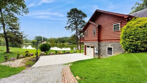 Une cabane en rondins à deux étages, avec des fondations en pierre, une allée pavée et un étang en arrière-plan.
