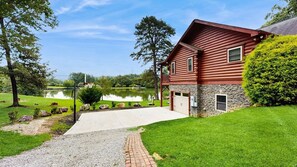 Eine zweistöckige Holzhütte mit Steinunterbau, einem gepflasterten Zufahrtsweg und einem Teich im Hintergrund.