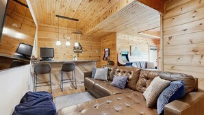 Un intérieur de chalet chaleureux avec un canapé en cuir, des tabourets de bar et un téléviseur.