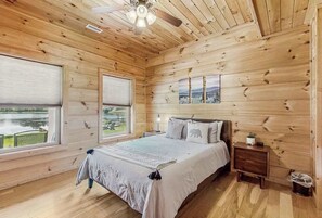 Ein Schlafzimmer einer Holzhütte mit einem Bett, Nachttischen, einem Deckenventilator und Blick auf einen See.
