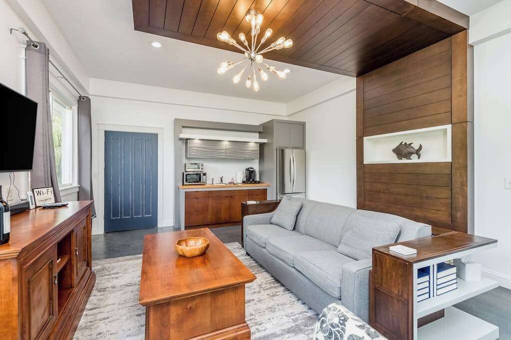A modern living room with a wooden coffee table, a grey sofa, a TV, and a kitchen area with a microwave and refrigerator.