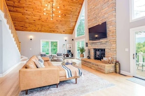 Ein geräumiges Wohnzimmer mit Holzbalkendecke, Kamin aus Stein, einem großen Fernseher und einer Couch.