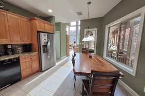 A kitchen with wooden cabinets, a stainless steel refrigerator, a dining table with chairs, and a large window with a view of the outdoors. - Mountain-View Escape on Rumbling Bald’s 14th & 15th Holes (Lake Lure)