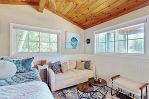 A cozy room with a bed, a sofa, a small table, and a bench. There are large windows with a view of trees.