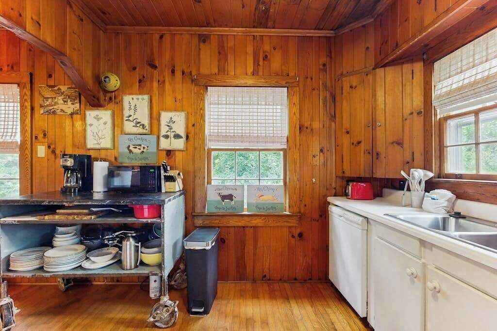 Eine Holzhütte mit einer Küche, die ebenfalls aus Holz gestaltet ist, einem Fenster mit Jalousien, einer Spüle und einer Mikrowelle.