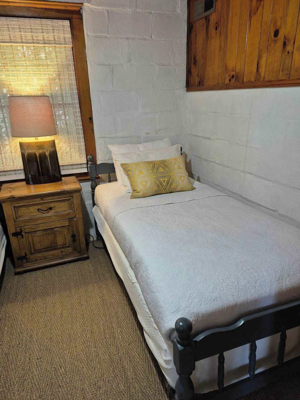 A bedroom with a bed, a nightstand, a lamp, a window with blinds, and a wooden wall panel.