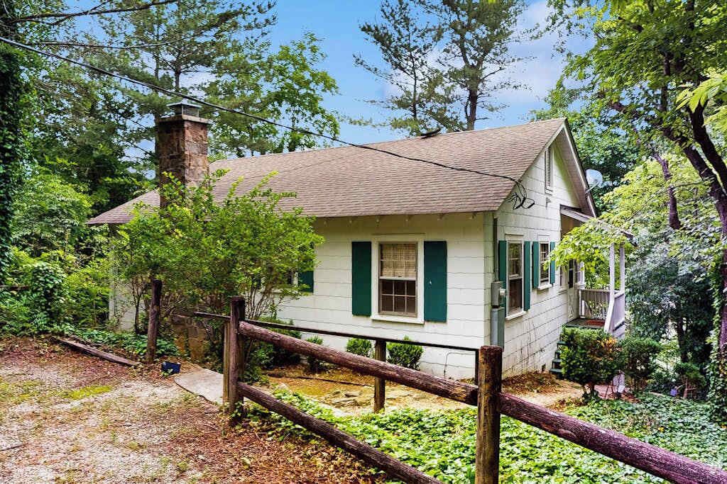 A small white house with green shutters, surrounded by trees and a wooden fence.