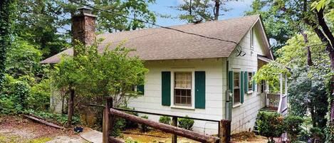 A small white house with green shutters, surrounded by trees and a wooden fence.