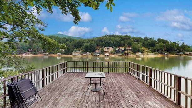 Une terrasse en bois avec une table et des chaises, donnant sur un lac paisible, avec des maisons en arrière-plan.