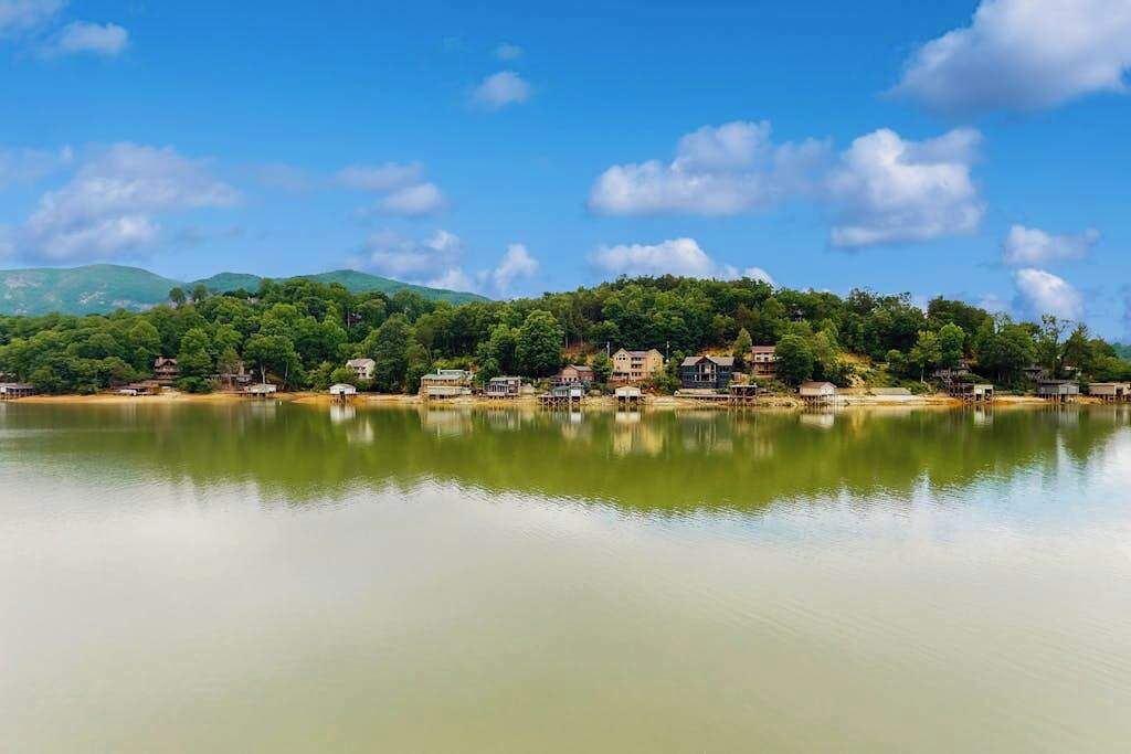 Blick auf einen See mit Gebäuden an einem Hang, ruhigem Wasser und blauem Himmel.