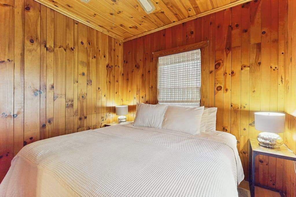A wooden-paneled room with a bed, two bedside tables, and a window with blinds.