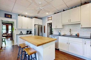 Eine Küche mit weißen Schränken, einer Holztischinsel mit Hockern, einem Deckenventilator und einem Edelstahlkühlschrank.