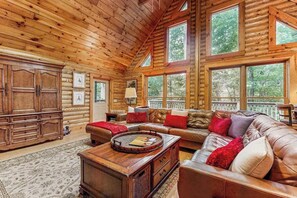 Ein rustikales Blockhaus-Interieur mit einer großen Couch, einem Holztisch und Blick auf den Wald durch große Fenster.