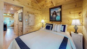 Um quarto estilo cabana de madeira, com uma cama, abajures, um quadro na parede e uma porta que dá para outro quarto.