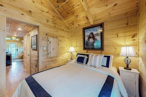 Ein holzgetäfeltes Zimmer im Chalet-Stil mit einem Bett, Nachttischlampen, einem gerahmten Bild an der Wand und einer Tür zu einem anderen Raum.