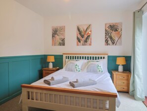A neatly made bed with white linens and two rolled towels, flanked by two bedside tables with lamps, in a room with teal walls and three tropical leaf wall art pieces. - 2 North Road (Dorchester)
