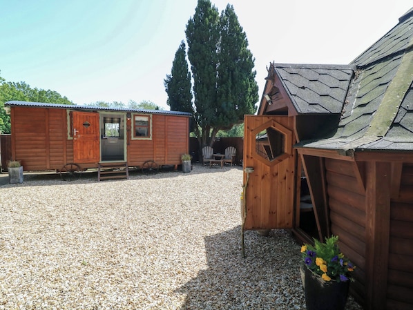A wooden cabin with a gravel driveway, a wheelbarrow, and a wooden structure with a hexagonal window. - Lake View Shepherds Hut (Lincoln)