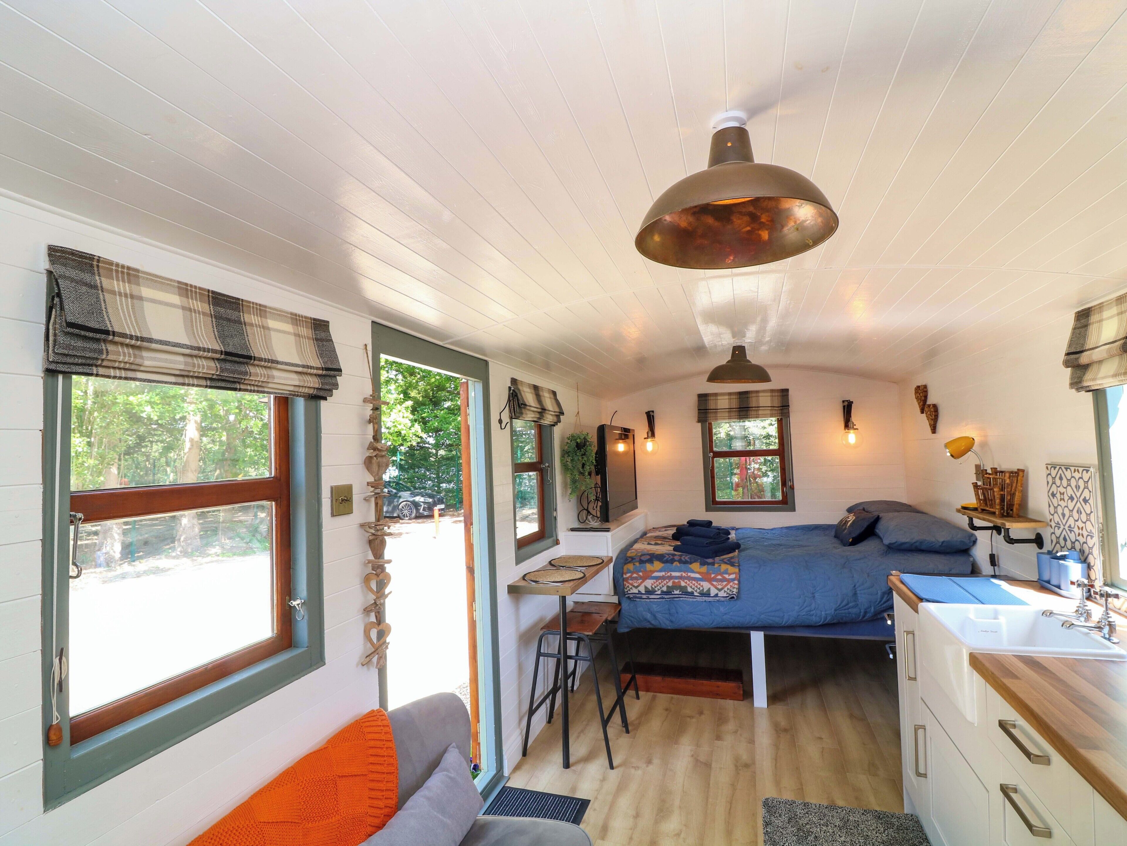 A compact room with a bed, desk, and kitchenette, featuring a large window and a hanging light fixture.