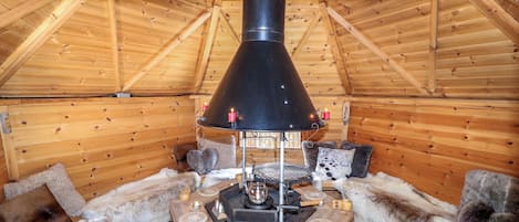 A cozy cabin interior with a wood stove, fur-covered seating, and a wooden ceiling.