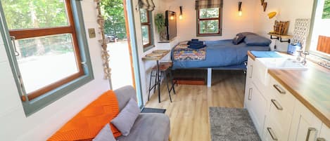 A compact RV interior with a bed, kitchenette, seating area, and large windows.