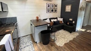 Una cucina compatta con un piano cottura moderno, una piccola zona pranzo con tavolo e sedie, un divano nero con cuscini a righe e un cestino nero.
