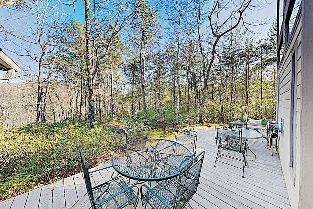 A deck with a glass-top table and metal chairs, surrounded by trees and a stone wall.
