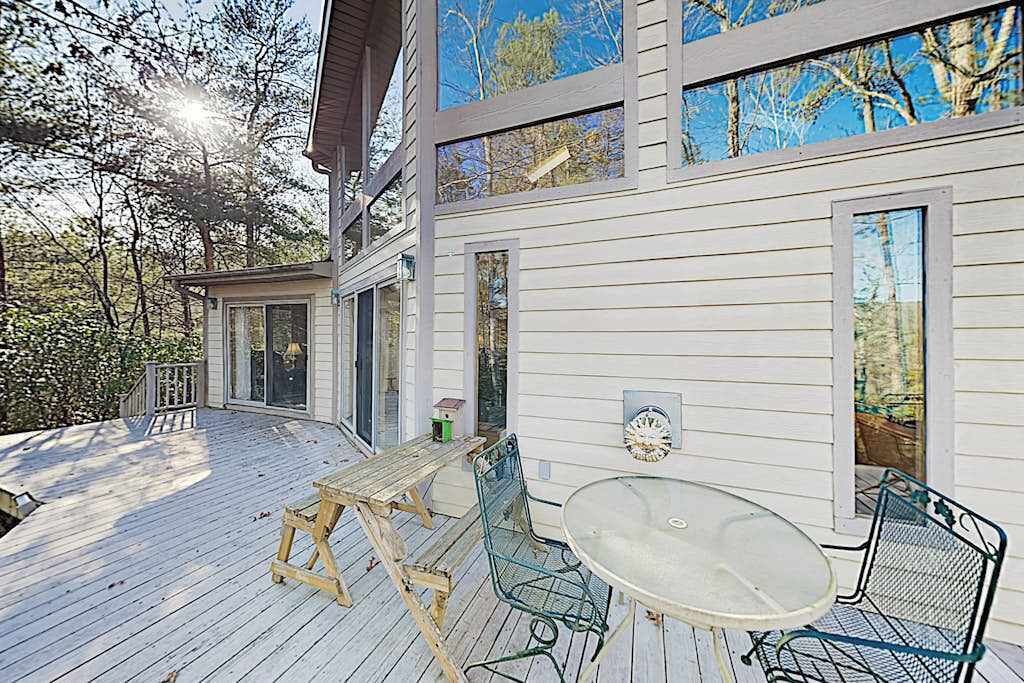 Eine Holzterrasse mit Tisch und Stühlen, ein überdachter Bereich mit einer Bank sowie ein Gebäude mit großen Fenstern.