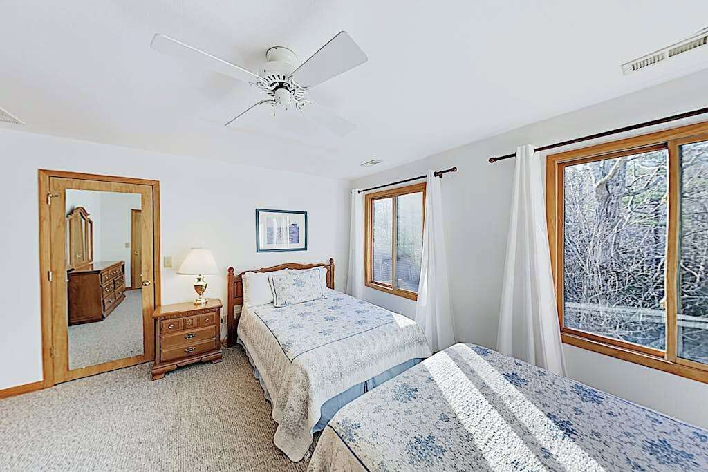 Une chambre à coucher comprenant un lit, une commode, un miroir, un ventilateur de plafond et une fenêtre donnant sur des arbres.