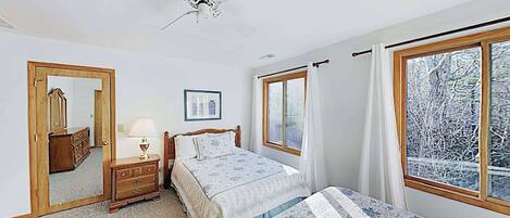 Une chambre à coucher comprenant un lit, une commode, un miroir, un ventilateur de plafond et une fenêtre donnant sur des arbres.