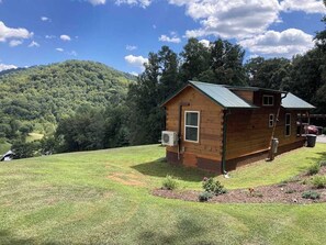 A wooden cabin with a metal roof, surrounded by a grassy area and trees, set against a mountainous backdrop. - Cozy cabin with amazing views, private hot tub, & covered porch. Nearby skiing. (Mars Hill)