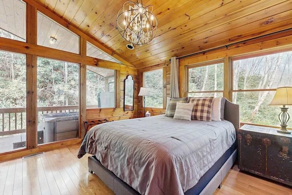 Un dormitorio de cabaña de madera con cama, un candelabro, ventanas grandes y vistas al bosque.