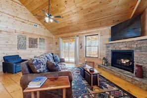 A cozy cabin interior with a wooden ceiling, a stone fireplace, a blue sofa, and a television mounted above the fireplace. - Highlands Water Hideaway (Murphy)