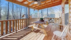 Eine Holzterrasse mit Tisch und Stühlen, einem Deckenventilator und Blick auf Bäume.