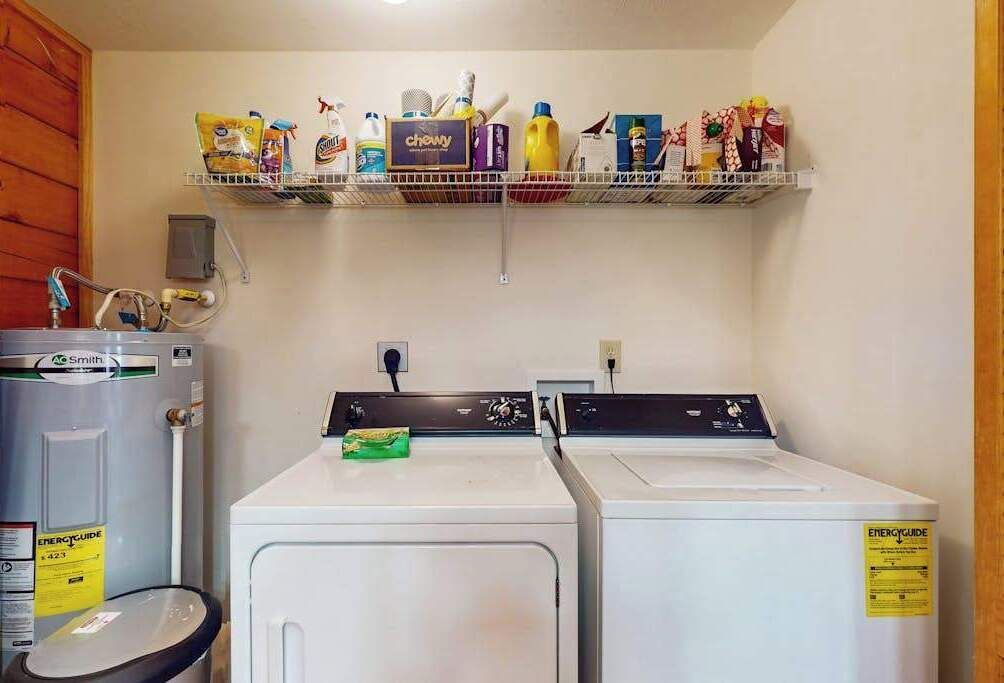 A laundry area with two white washers and dryers, a metal shelf above with cleaning supplies, and a water heater to the left.
