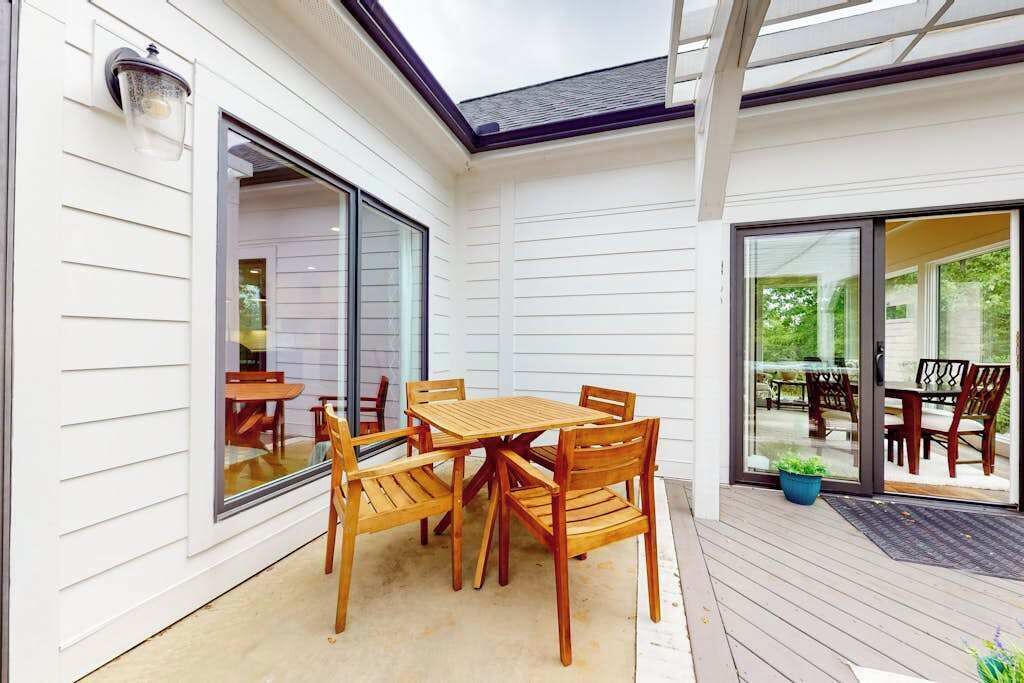 Eine überdachte Terrasse mit Holztisch und -stühlen, großen Schiebeglastüren und einem Blumentopf.