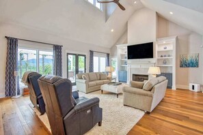A spacious living room with a fireplace, a flat-screen TV, and a large window with curtains. - Mountain & lake-view luxury home with hot tub, deck, & pergola - dog-friendly (Mill Spring)
