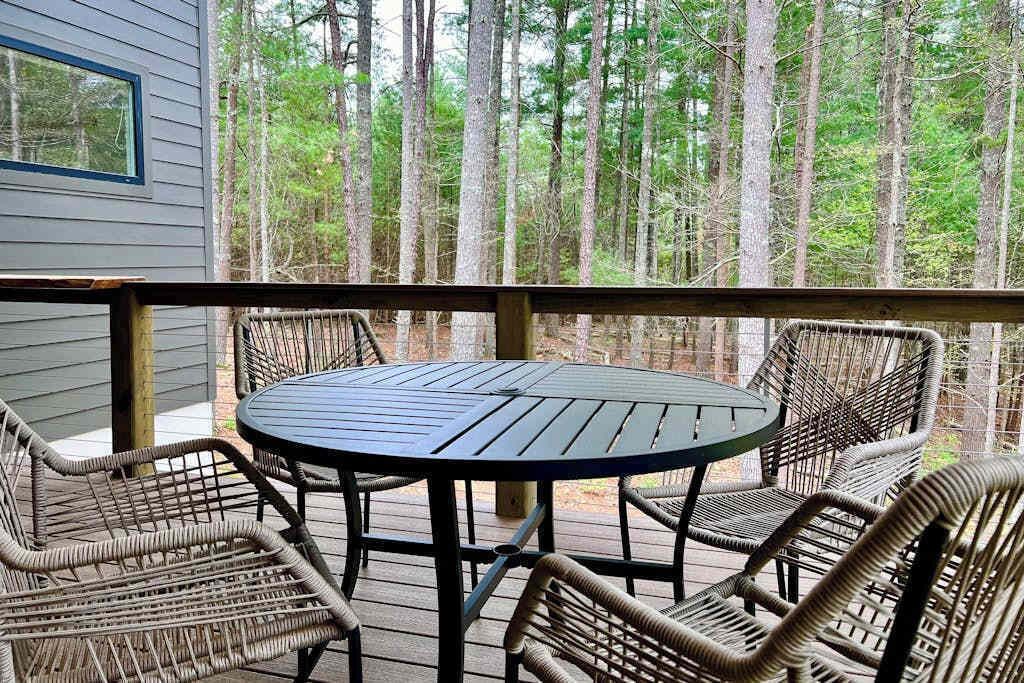 A covered outdoor area with a round table and wicker chairs, surrounded by a wooden deck and overlooking a forest.