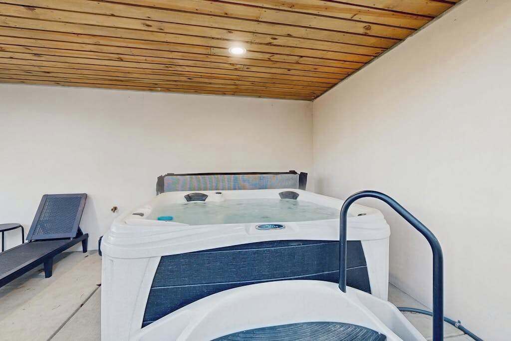 A hot tub with a metal frame, a lounge chair, and a small table in a room with wooden ceiling and white walls.